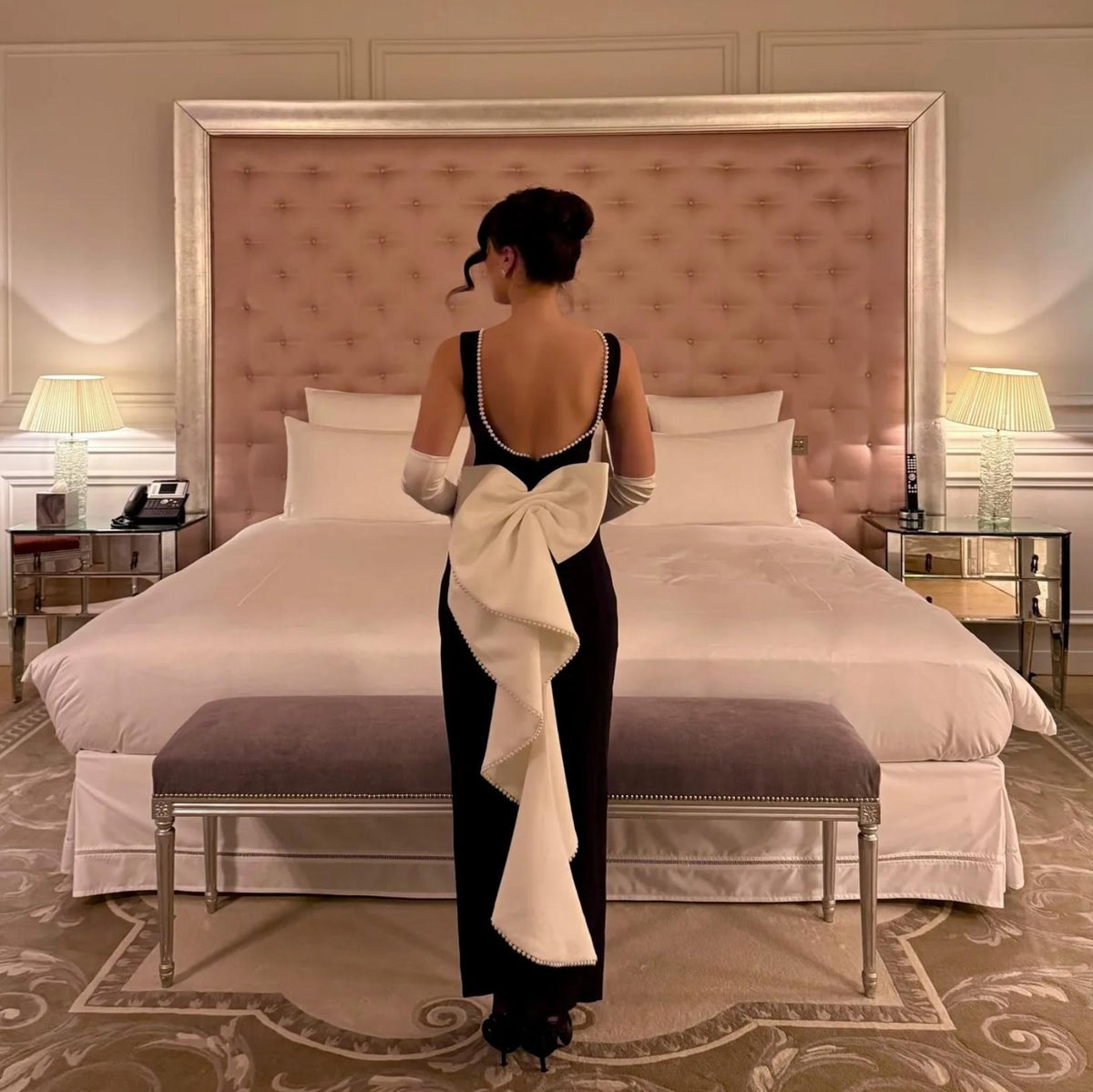Black satin midi dress with a large white back bow, pearl trim, and elegant silhouette in a luxury hotel room.
