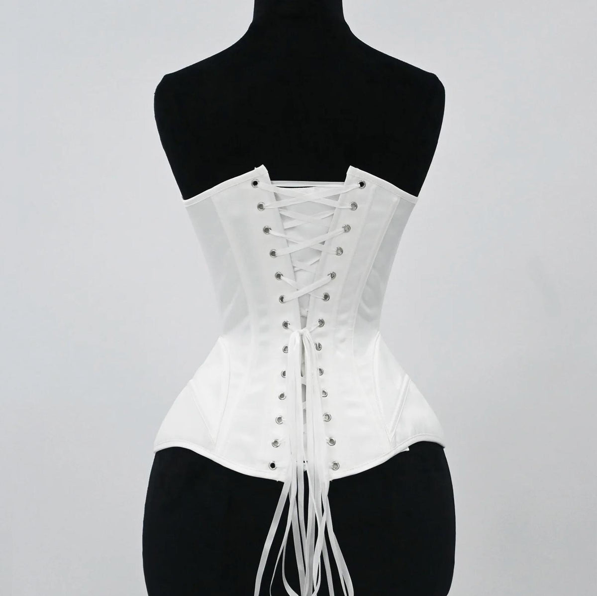 White satin bridal corset featuring a structured lace-up back with silver grommets and long ribbon ties on a mannequin.