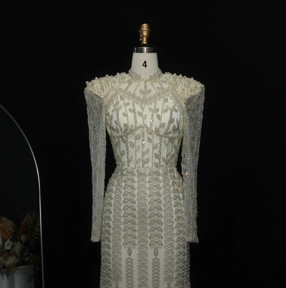 White mermaid gown featuring intricate crystal and pearl beading, long sleeves, and a high neck on a mannequin.