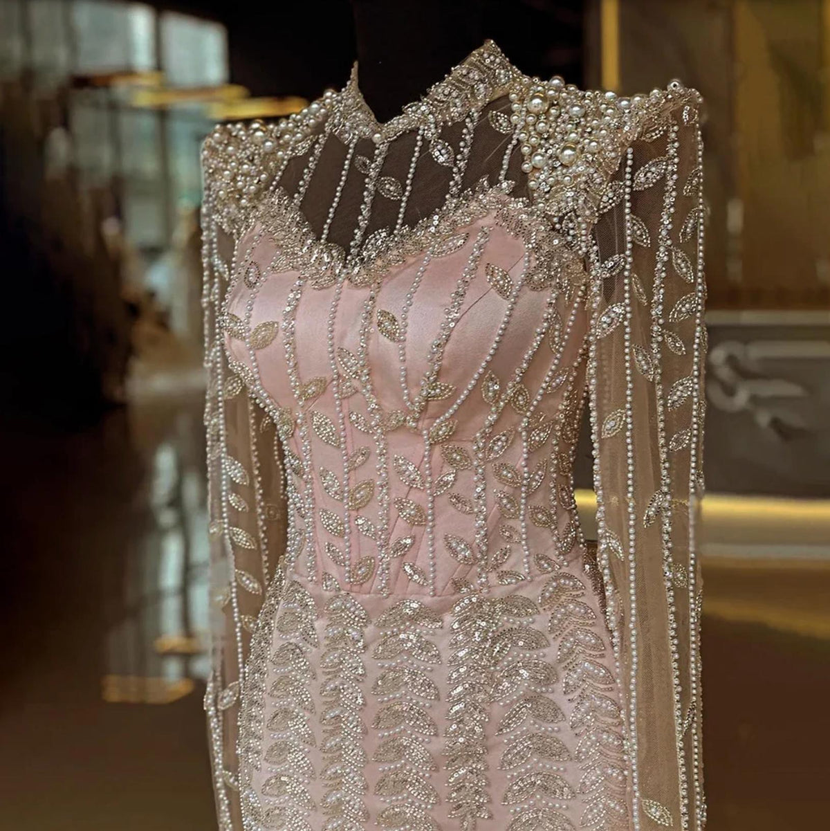 Blush pink mermaid gown featuring intricate pearl and crystal beading, sheer tulle sleeves, and a high-neck bodice.