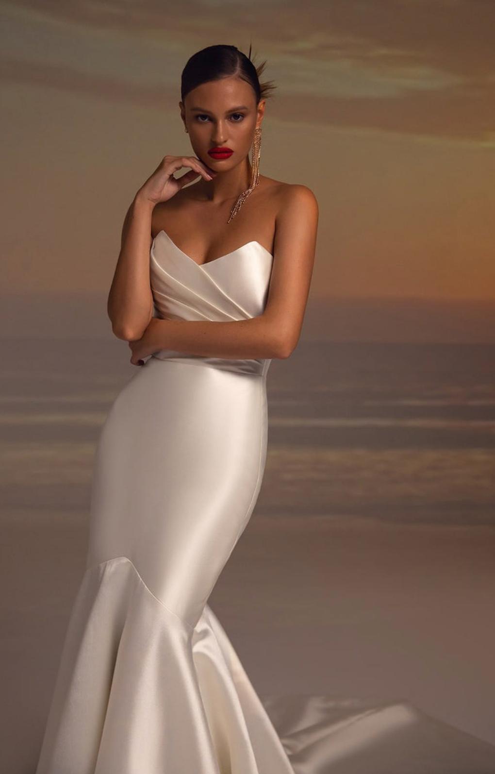 Elegant white satin fishtail gown with pleated bodice detailing, worn by a model against a sunset beach backdrop.