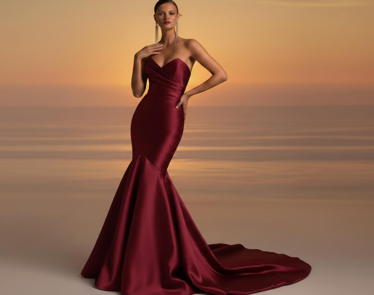 Burgundy satin fishtail gown with a strapless sweetheart neckline and pleated bodice against a sunset background.