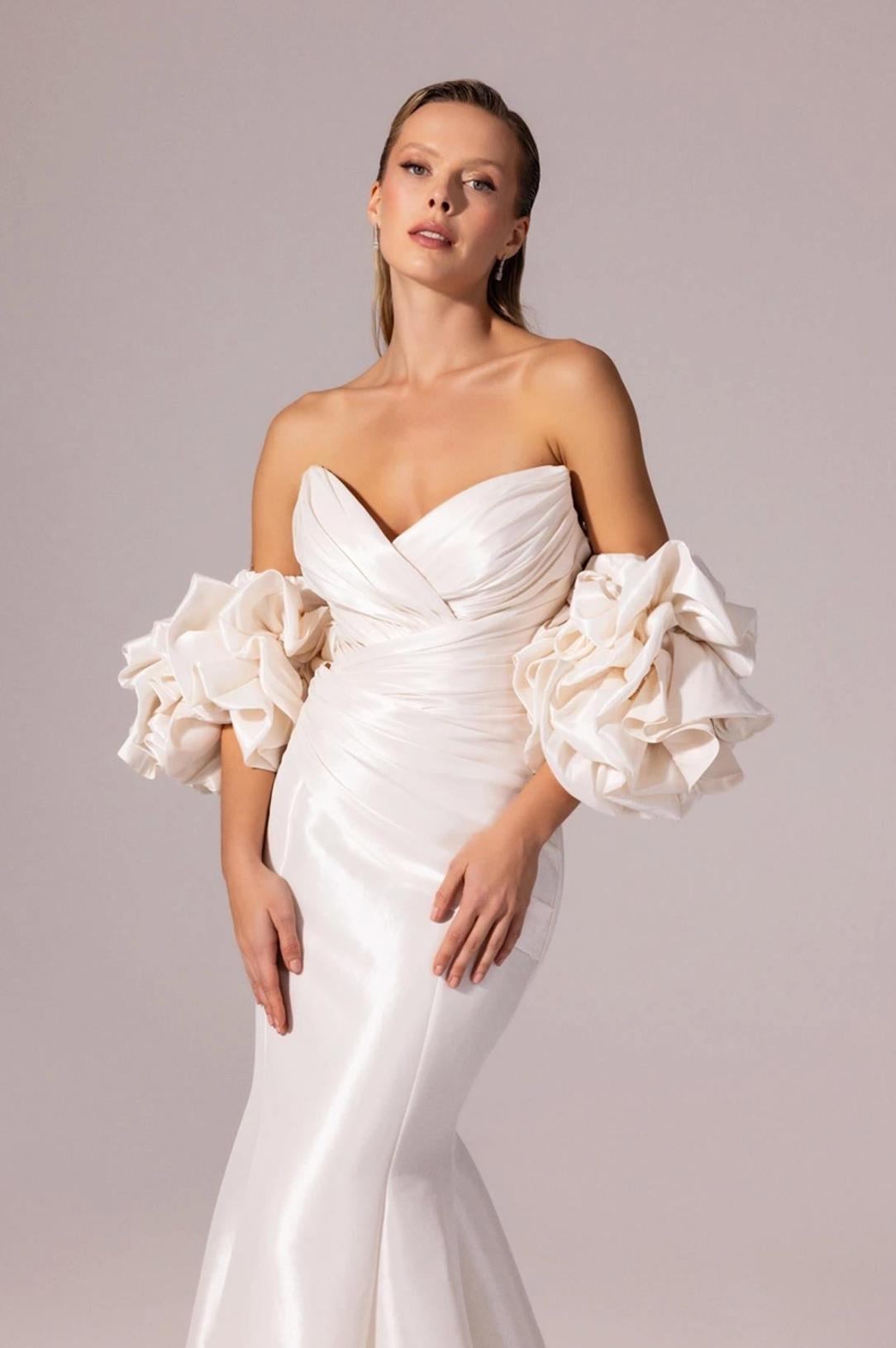 White satin mermaid gown with a sweetheart neckline and dramatic, voluminous ruffled off-the-shoulder sleeves.
