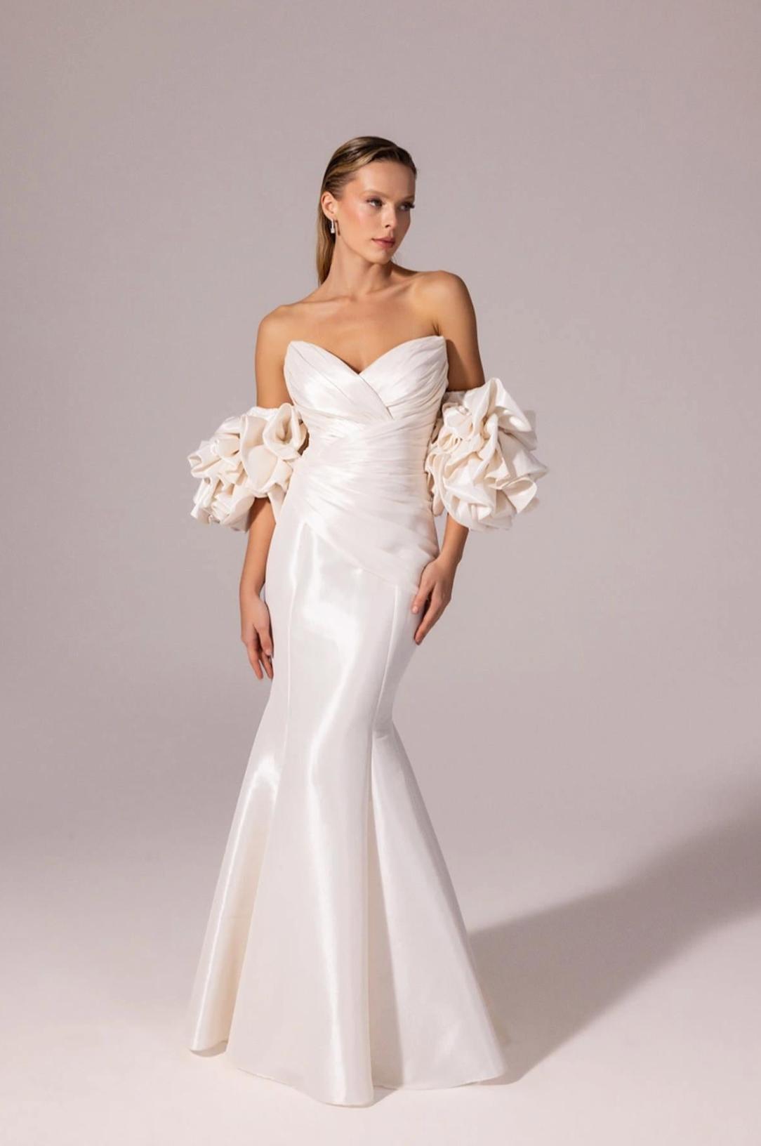 White satin mermaid gown with a sweetheart neckline and dramatic ruffled off-the-shoulder sleeves.
