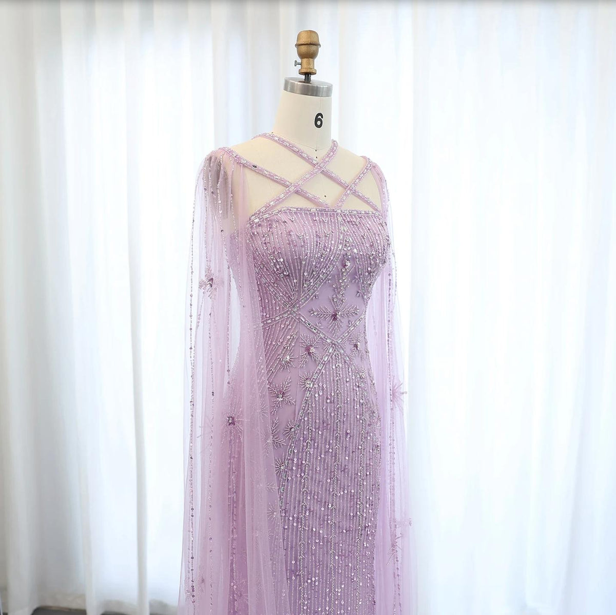 Lavender mermaid gown with tulle cape sleeves, intricate crystal beading, and a criss-cross neckline on a mannequin.