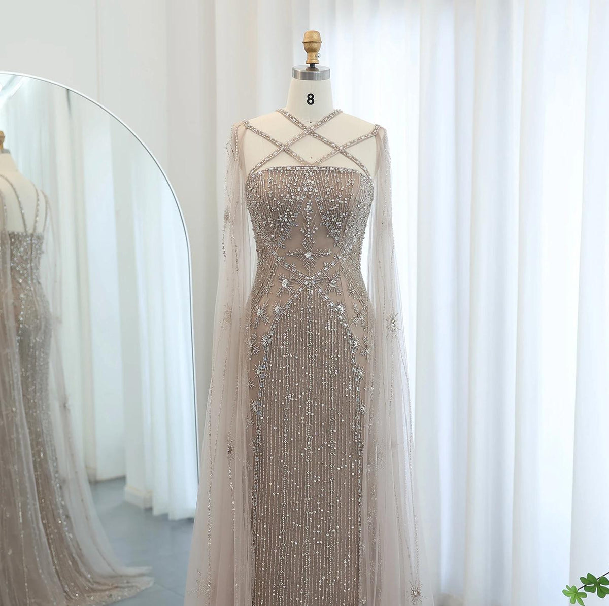 Beaded mermaid gown with tulle cape sleeves, criss-cross neckline, and shimmering crystal embellishments on a mannequin.