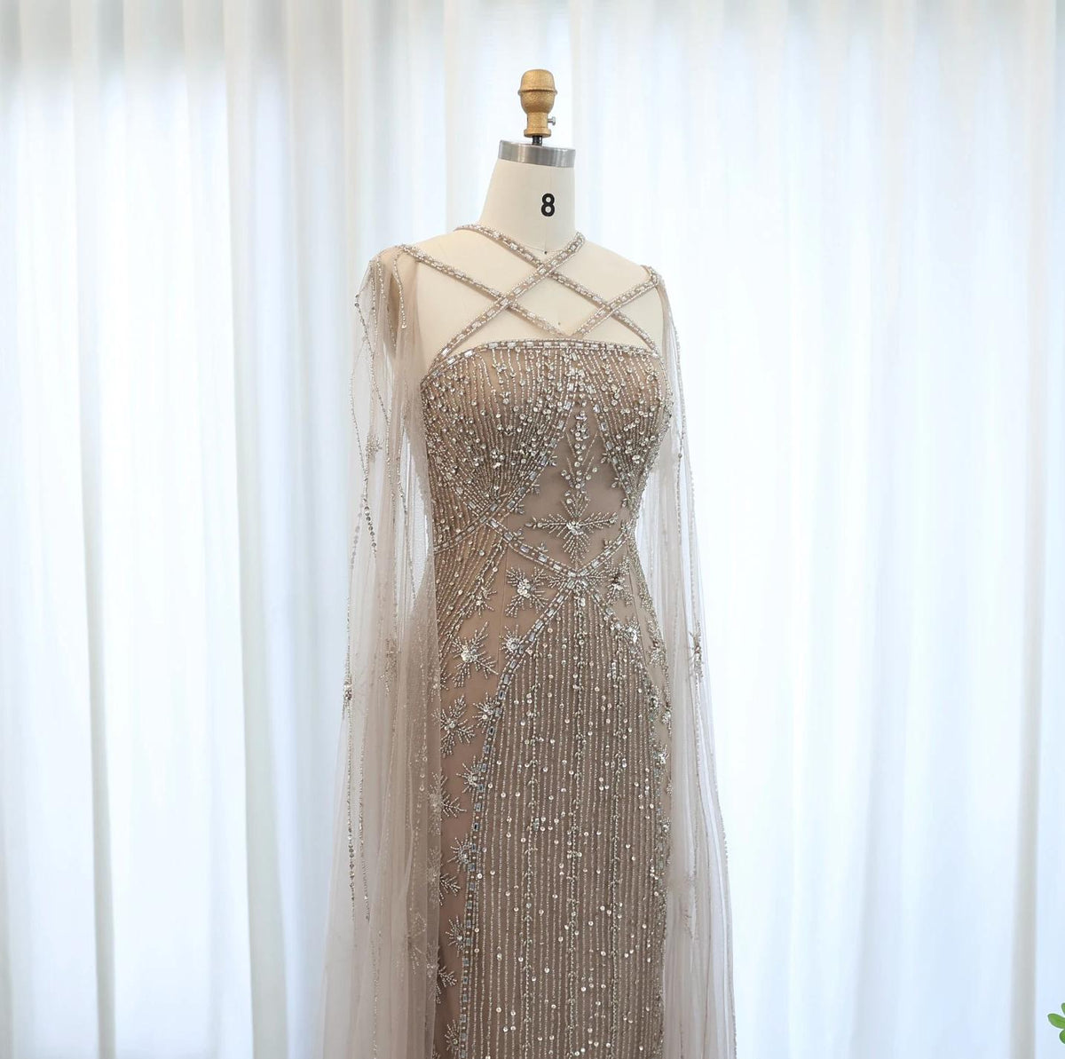 Beaded mermaid gown with criss-cross neckline, shimmering crystals, and sheer tulle cape sleeves on a mannequin.