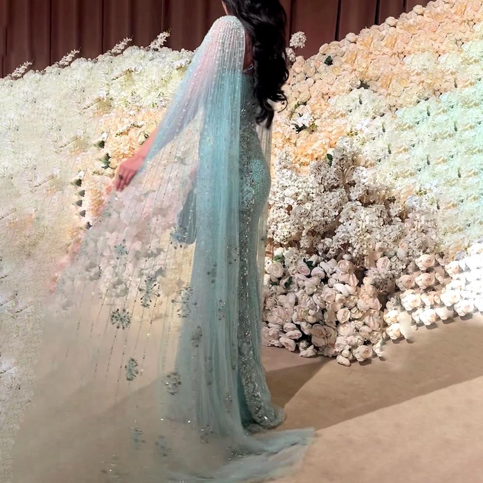 Light blue mermaid gown with dramatic tulle cape sleeves, intricate beading, and shimmering sequins against a floral wall.