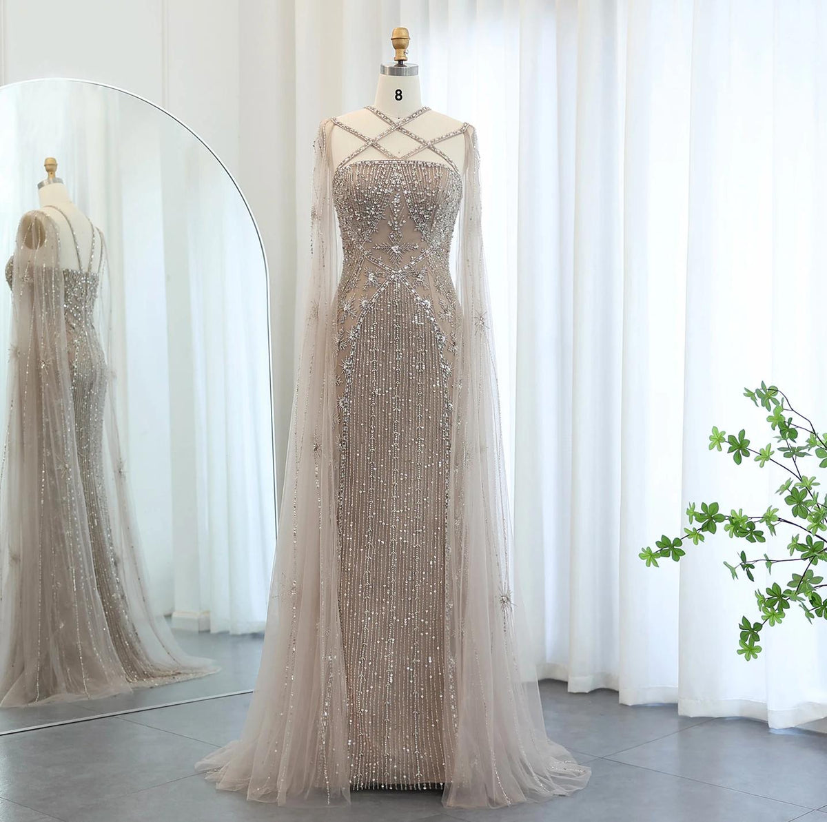 Beaded mermaid gown with tulle cape sleeves, criss-cross neckline, and shimmering crystal embellishments on a mannequin.