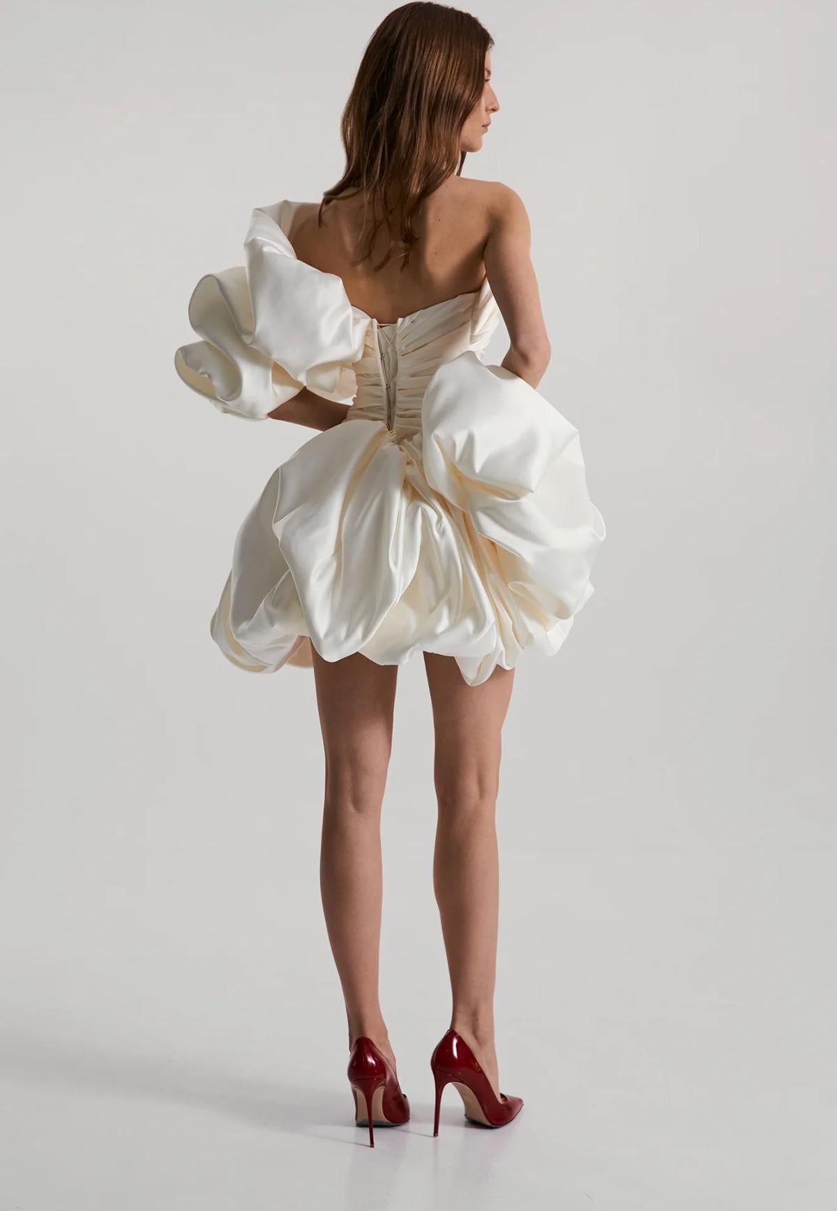 Back view of a white strapless mini dress in ruched satin with a voluminous ruffled skirt and lace-up back detail.
