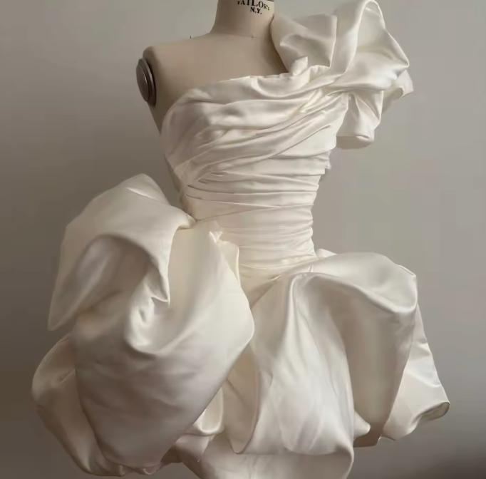 White satin mini dress with a ruched bodice, one-shoulder ruffled neckline, and voluminous skirt on a mannequin.