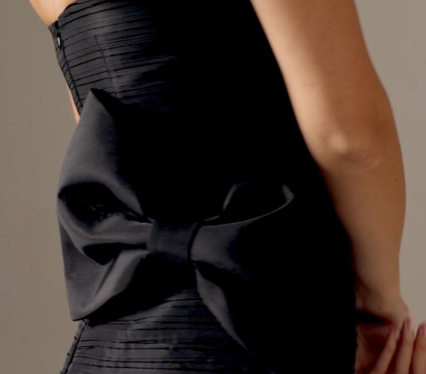 Back view of a black strapless satin Candelaria gown featuring a dramatic oversized bow and elegant pleated bodice details.