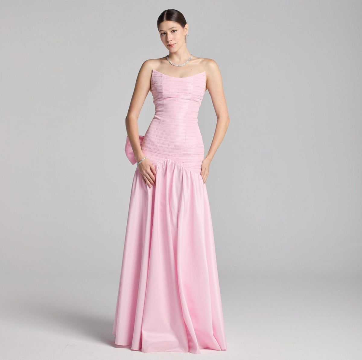 Model wearing a strapless pink satin A-line Candelaria gown with pleated bodice detailing and a dramatic back bow.