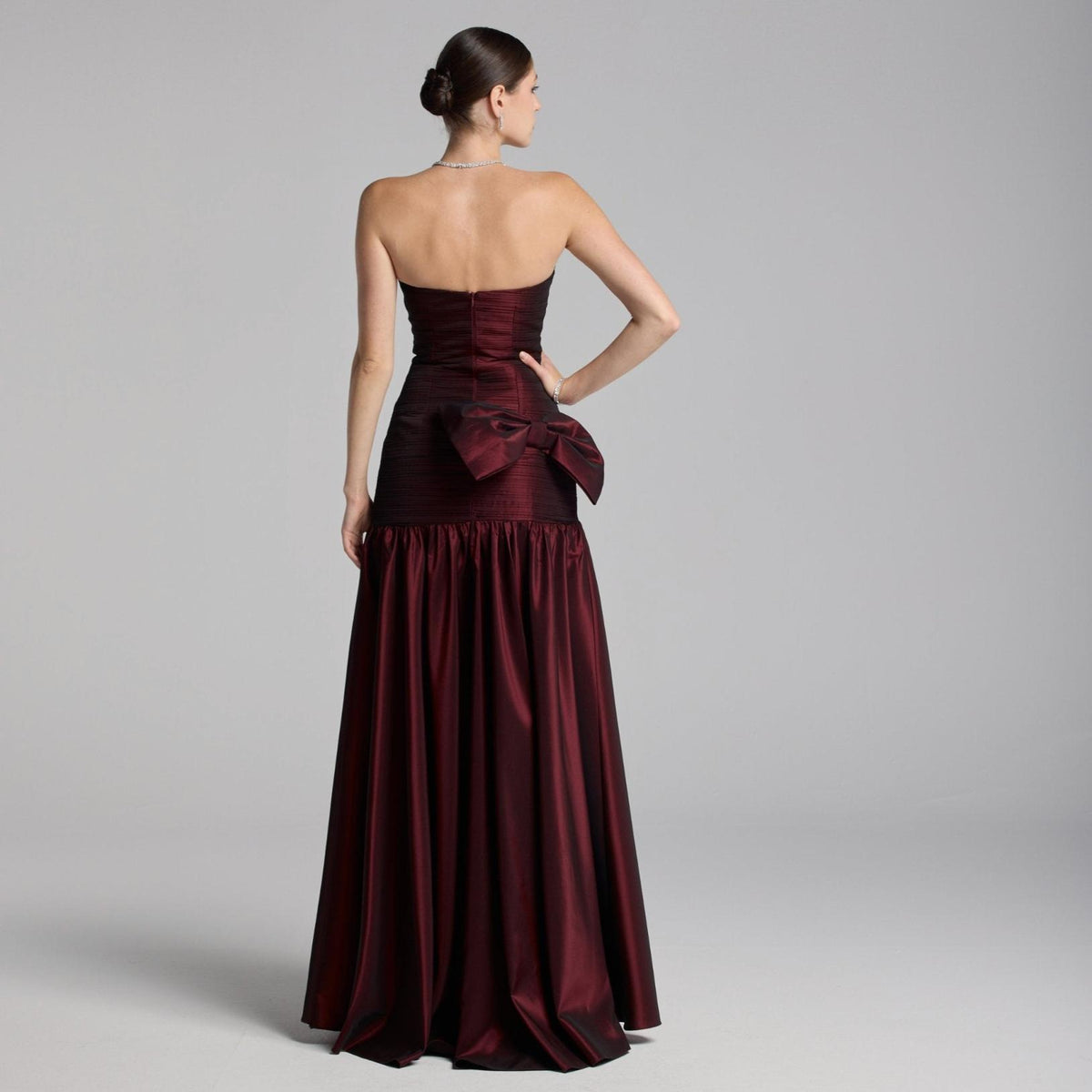Back view of a strapless burgundy satin A-line gown featuring a dramatic back bow and pleated bodice details.