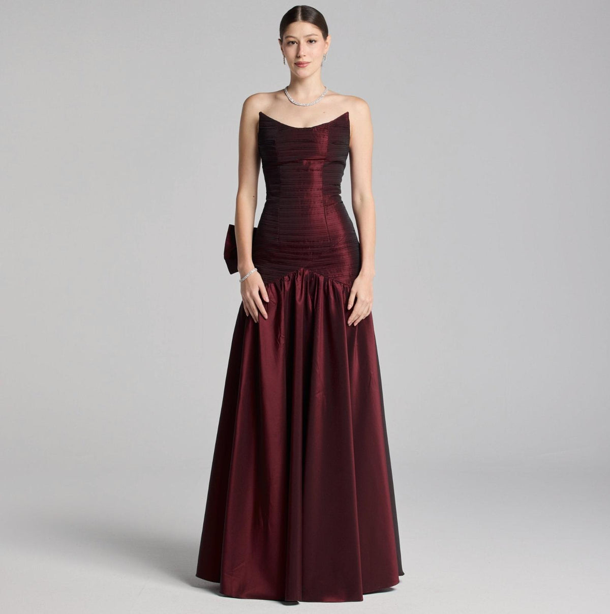 Strapless burgundy satin A-line evening gown with pleated bodice and dramatic back bow detail.