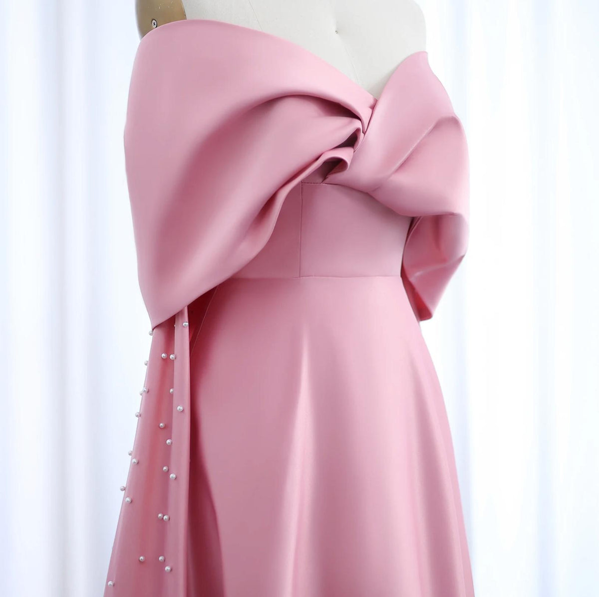 Dubai pink satin off-shoulder gown with cap sleeves, draped bodice, and delicate pearl embellishments on a mannequin.