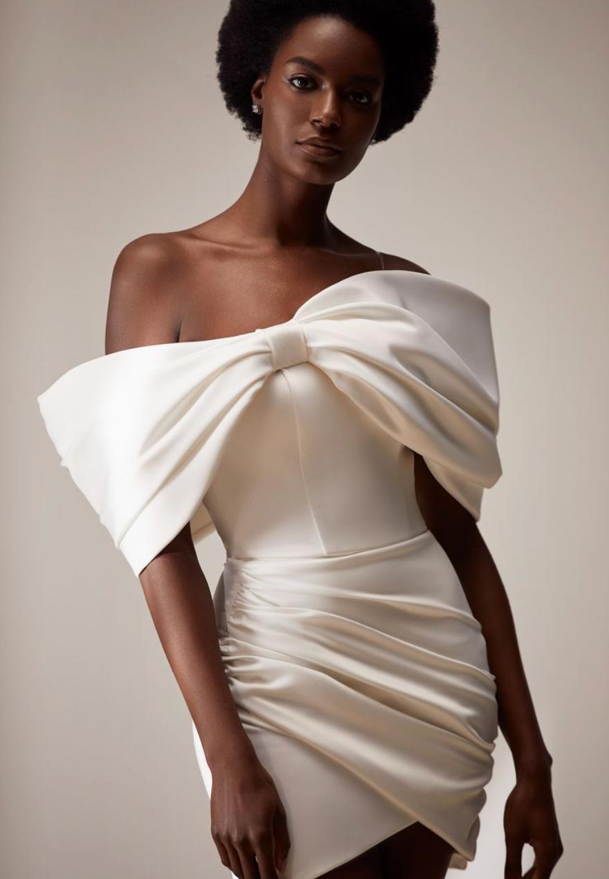 Off-the-shoulder white satin mini dress with a ruched bodice and dramatic draped bow detail.