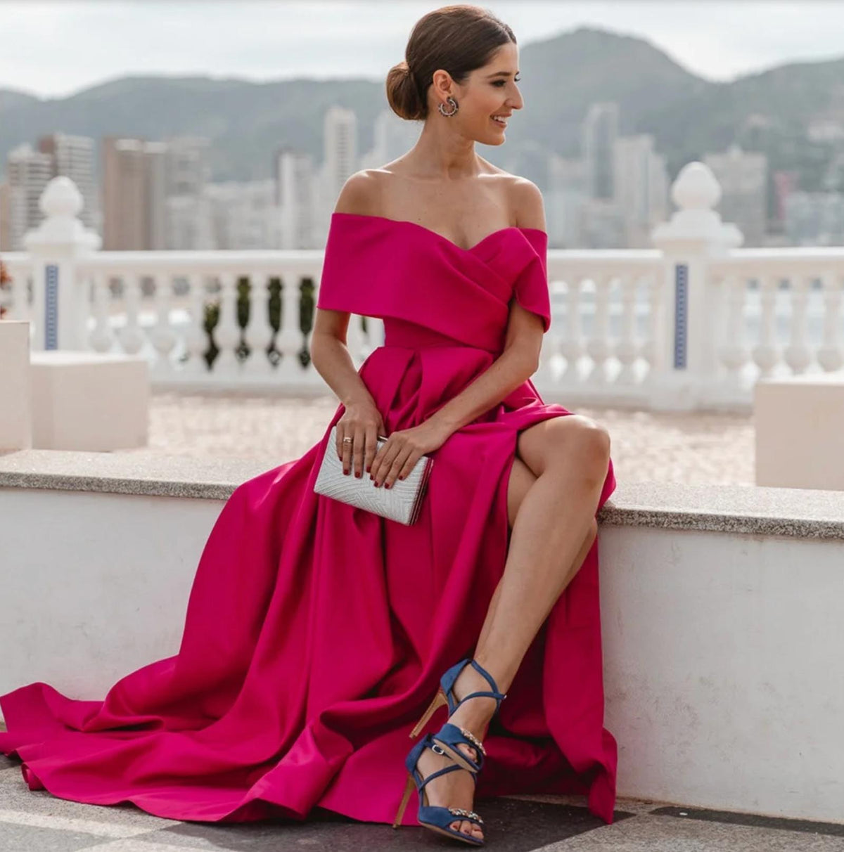 Fuchsia satin A-line gown with an off-shoulder neckline and side slit, worn with blue heels and a silver clutch.