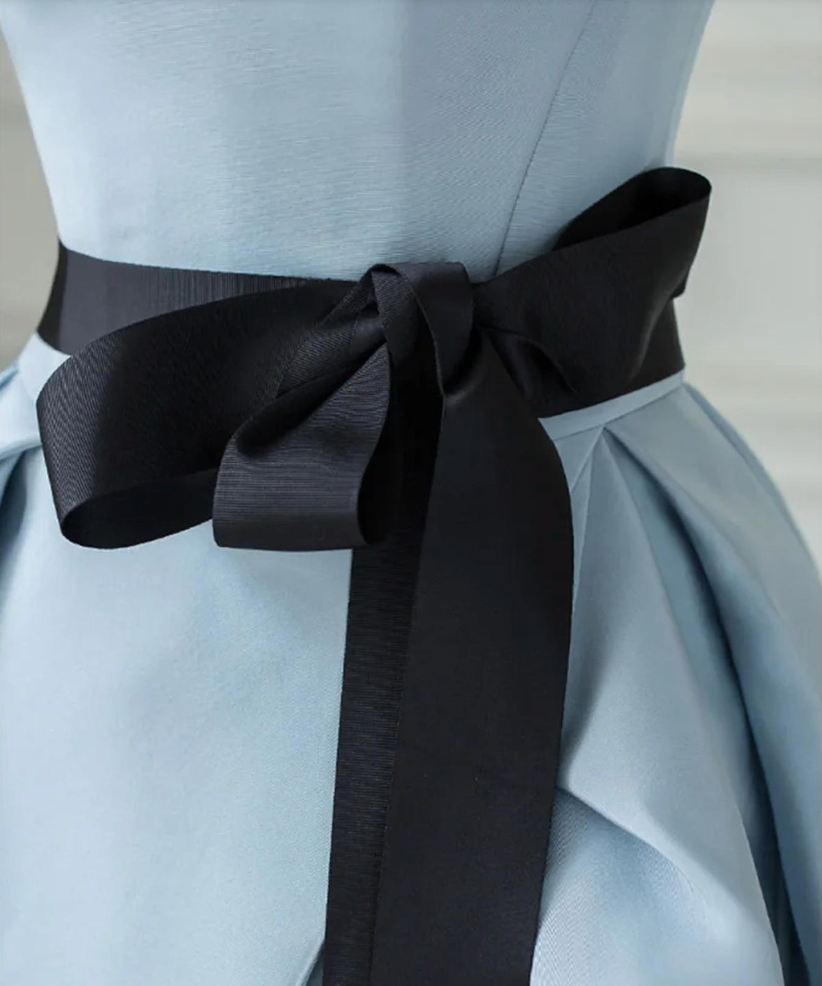 Light blue satin twill Crete gown featuring a pleated silhouette and a contrasting black sash tied at the waist.
