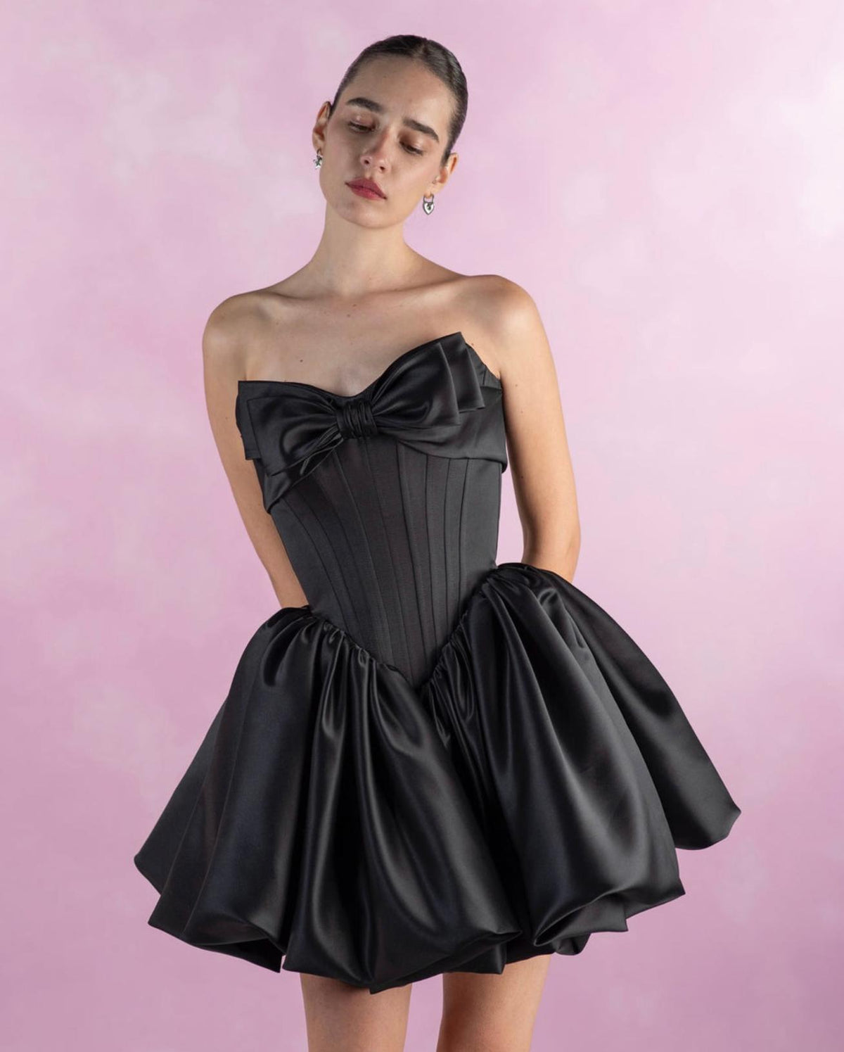 Strapless black satin mini dress with a structured bodice, large front bow, and voluminous pleated A-line skirt.