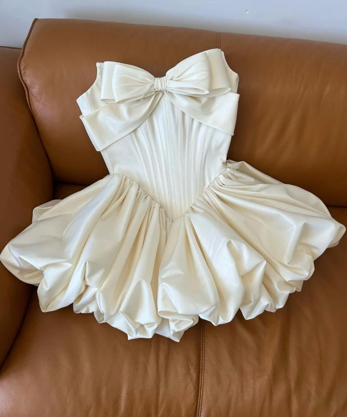 Ivory satin mini bridal dress with a strapless bodice, large front bow, and a voluminous pleated A-line skirt.