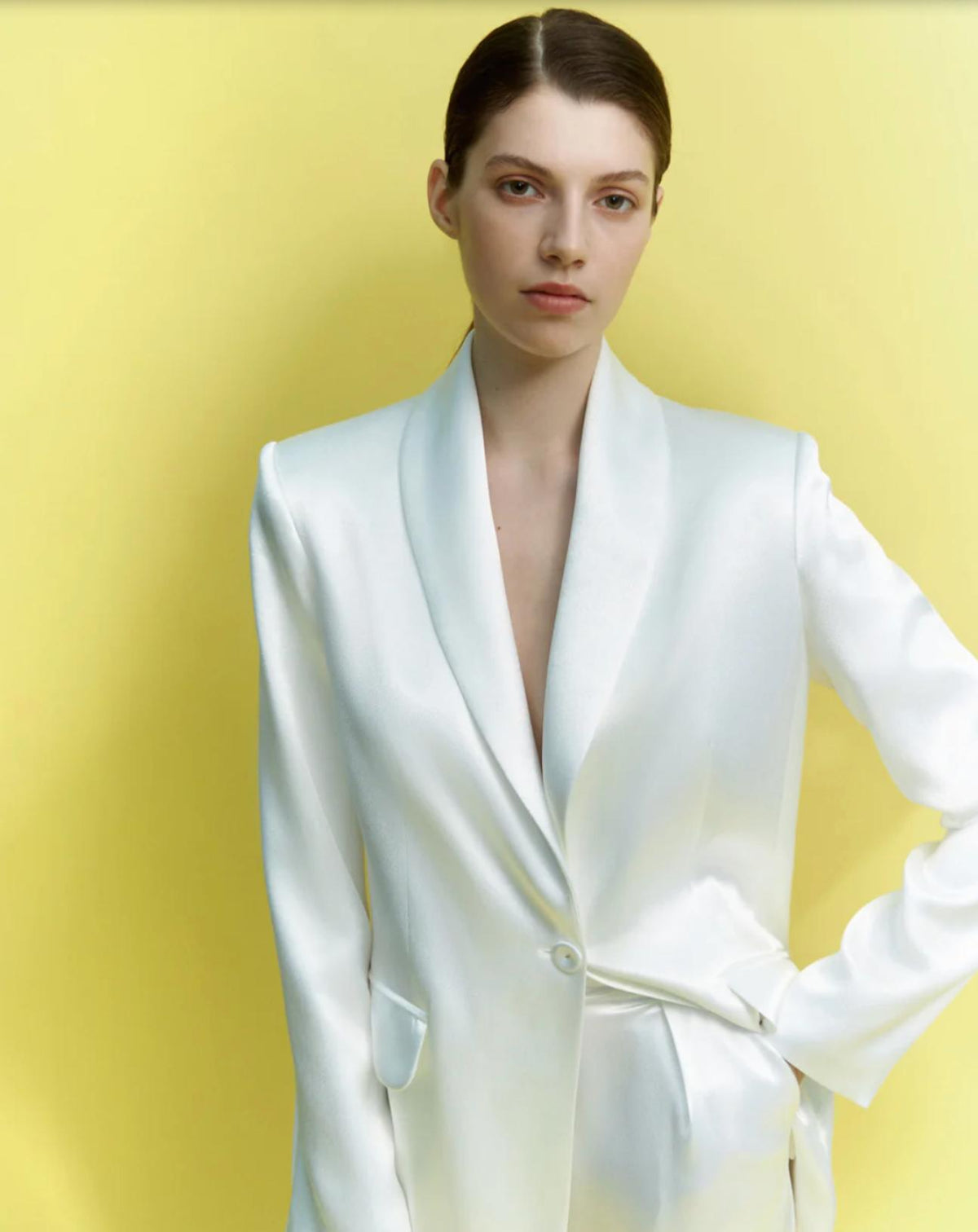 Woman wearing a white satin two-piece suit with a single-button shawl lapel jacket against a yellow background.
