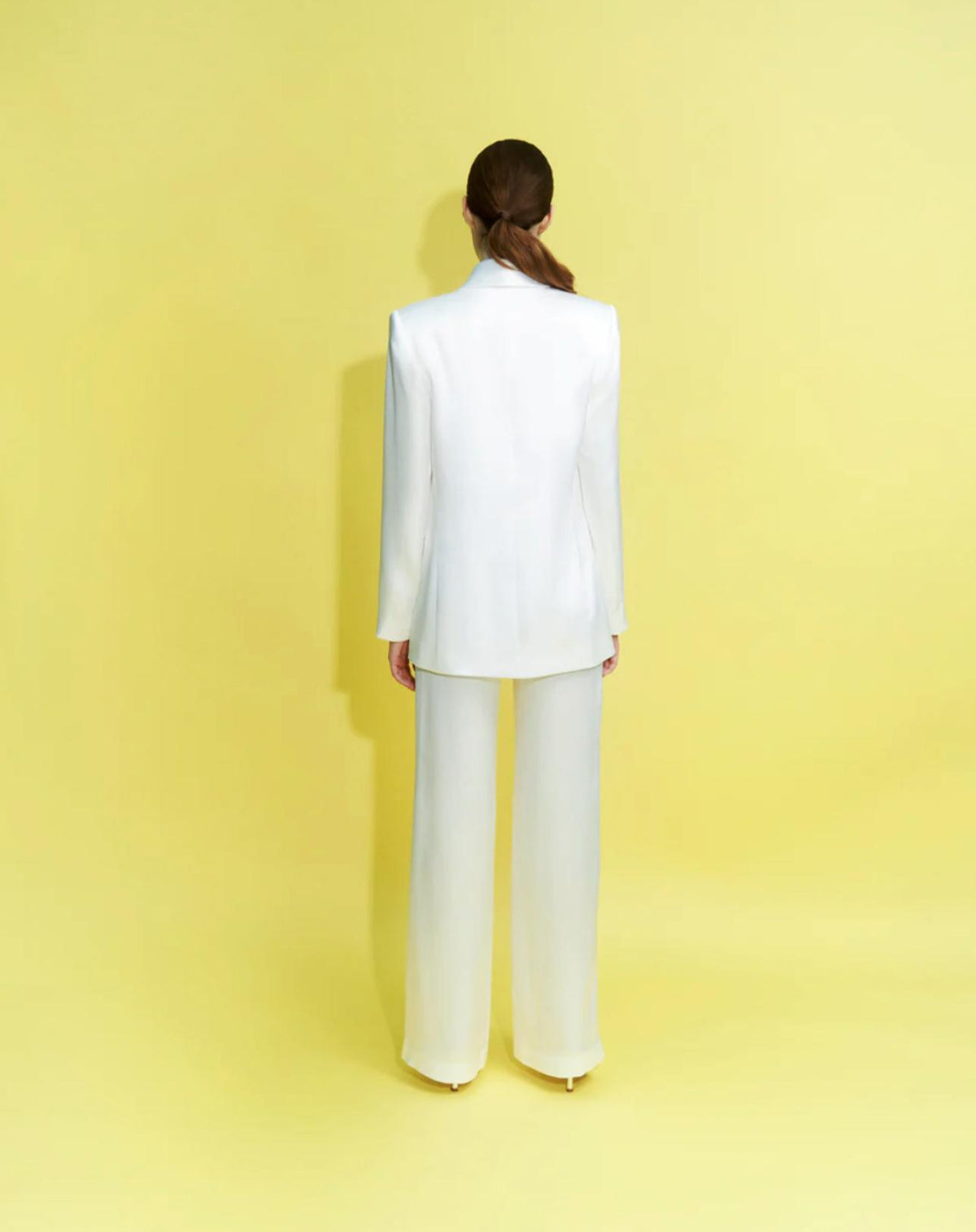 Back view of a woman wearing a white satin two-piece suit with a tailored shawl lapel jacket and wide-leg trousers.