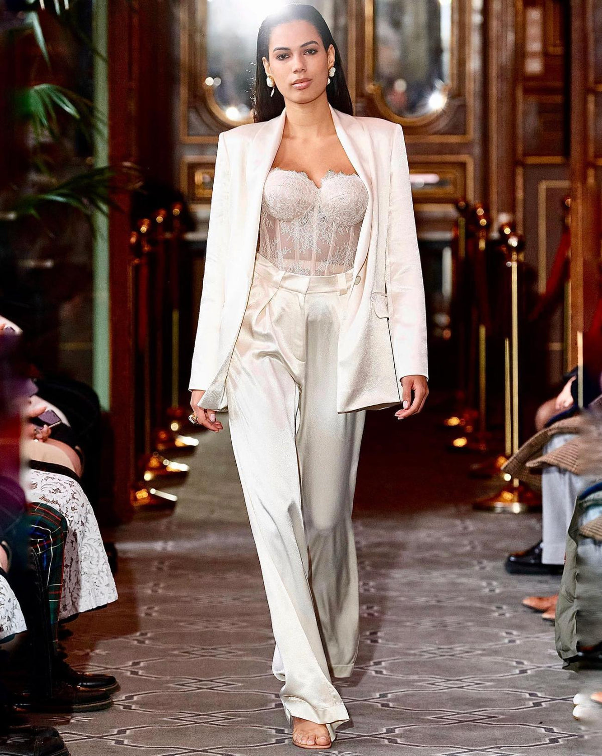 Model wearing a cream satin two-piece suit with a shawl lapel jacket, wide-leg trousers, and a lace corset top on a runway.