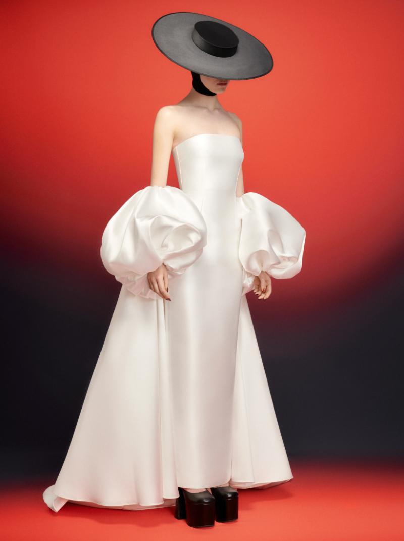 White satin strapless evening gown with dramatic puff sleeve cape, worn with a wide-brimmed black hat against a red background.