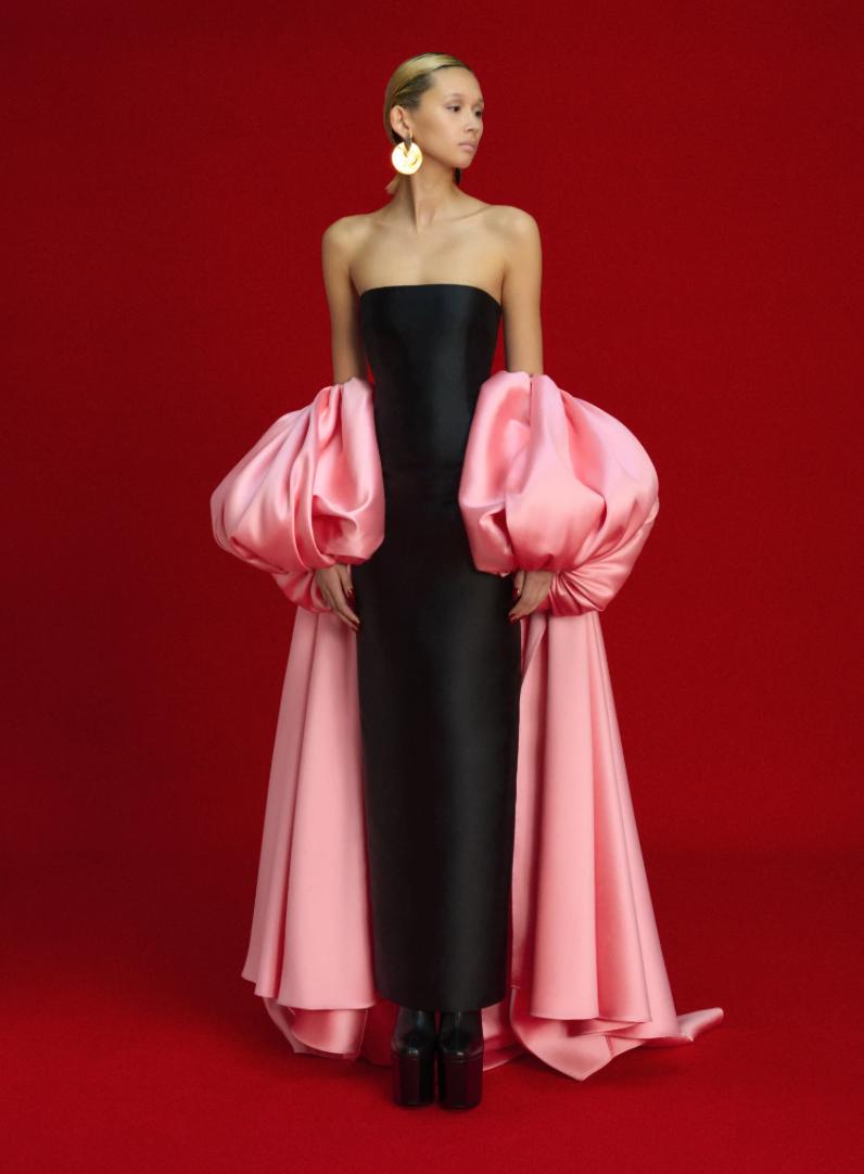 Strapless black satin evening gown featuring a dramatic, removable pink puff sleeve cape against a red background.