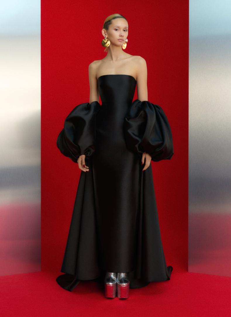 Black strapless satin evening gown with dramatic puff sleeves and a floor-length skirt against a red background.