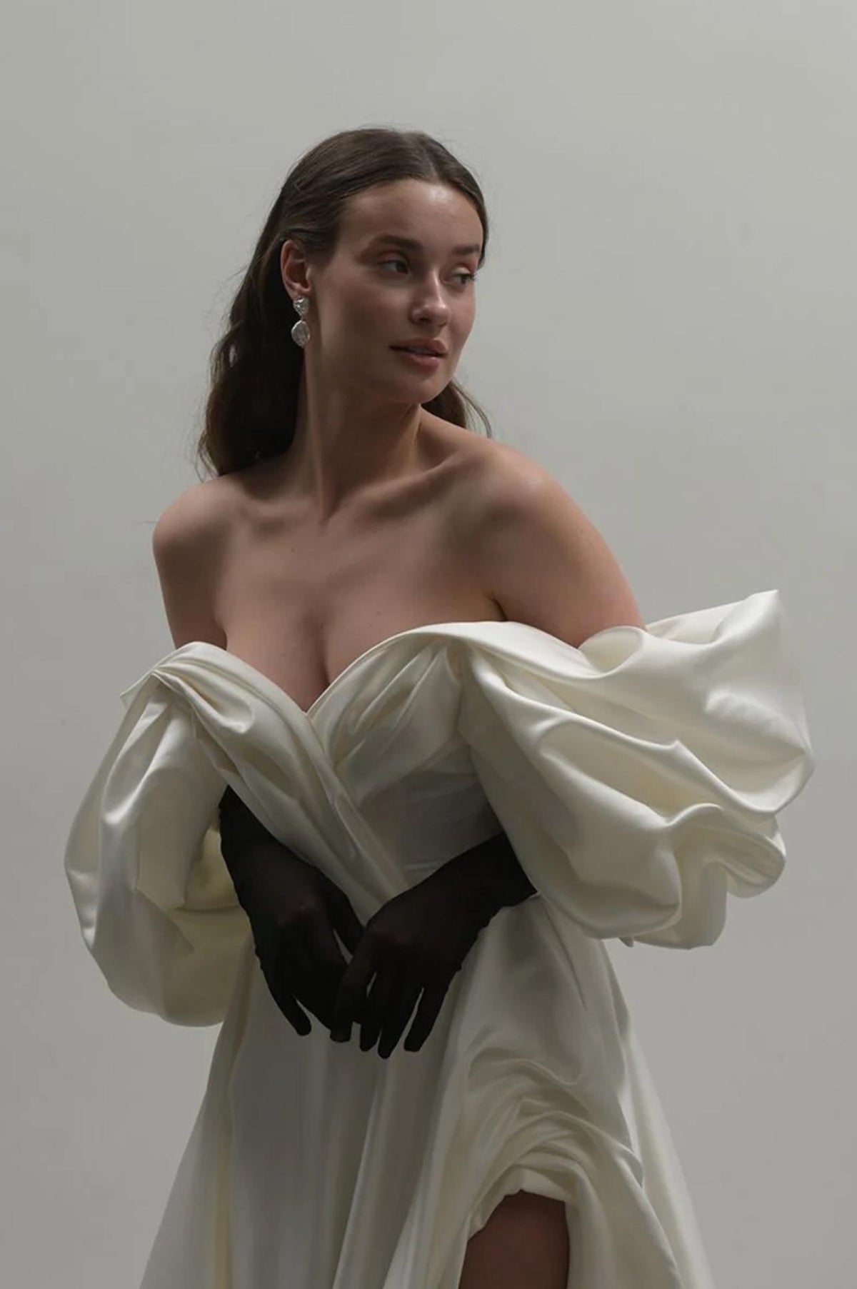 Off-shoulder white satin A-line gown with voluminous pleated sleeves and a high side slit, styled with black gloves.