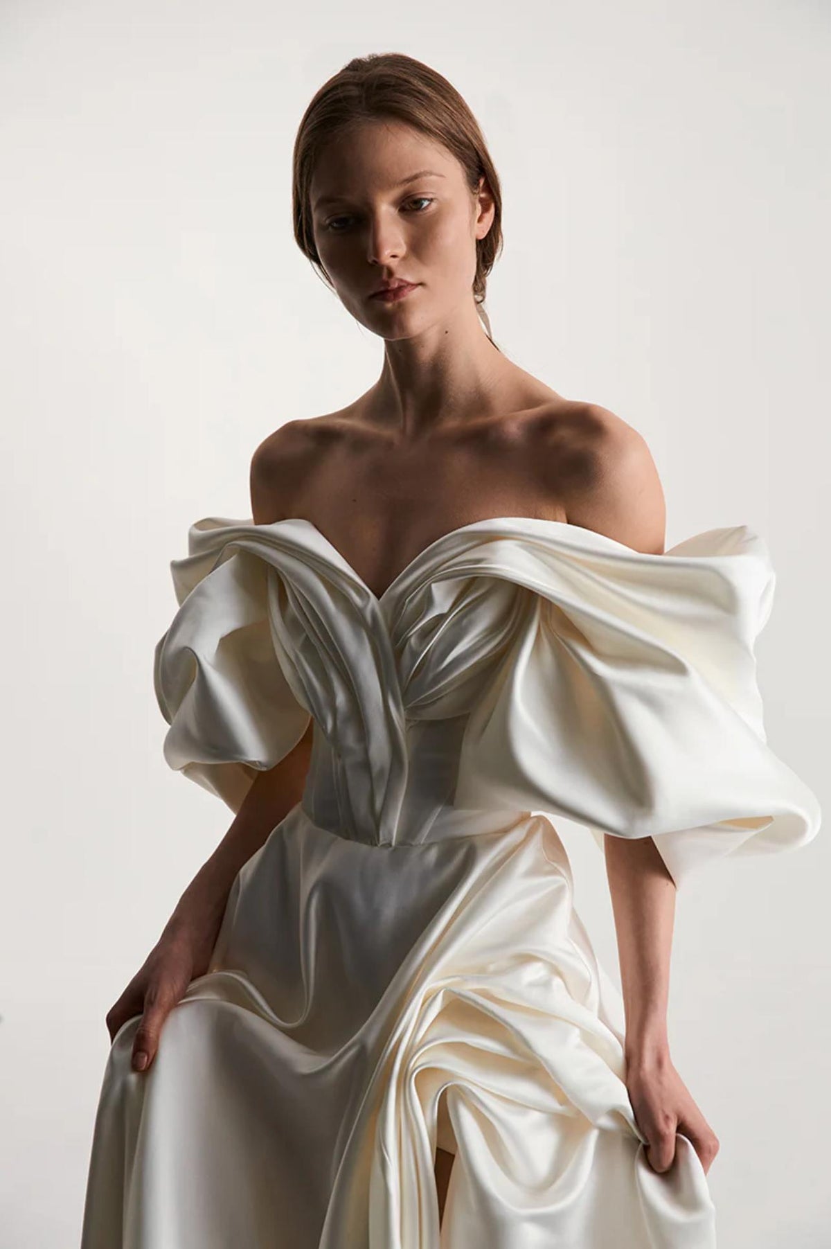 Off-shoulder ivory satin A-line gown featuring dramatic pleated sleeves and a structured bodice on a model.