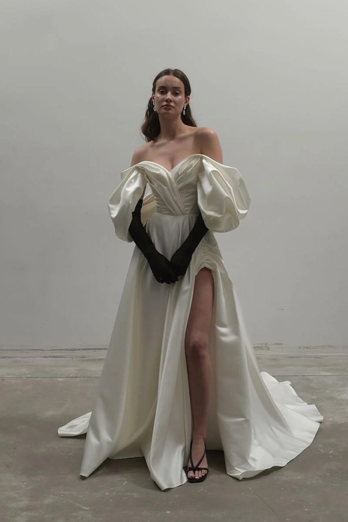 Off-shoulder white satin A-line gown with voluminous sleeves, a high side slit, and elegant black opera gloves.