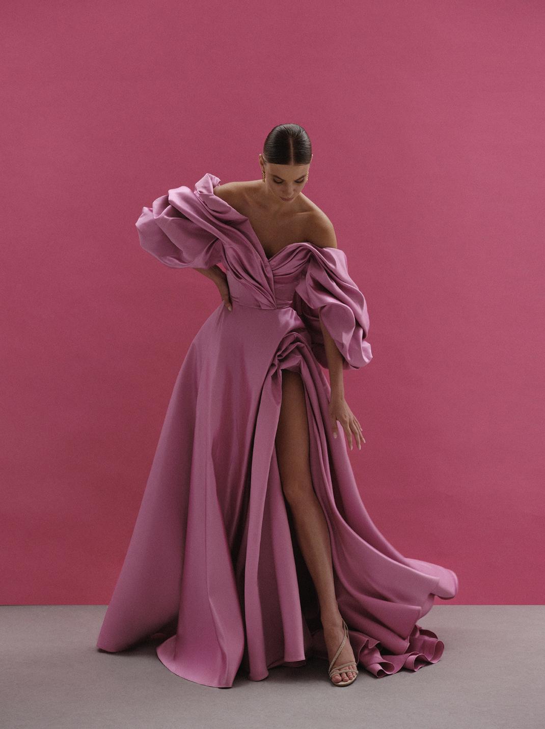 Pink satin off-shoulder A-line gown with voluminous sleeves, pleated bodice, and a daring high side slit.