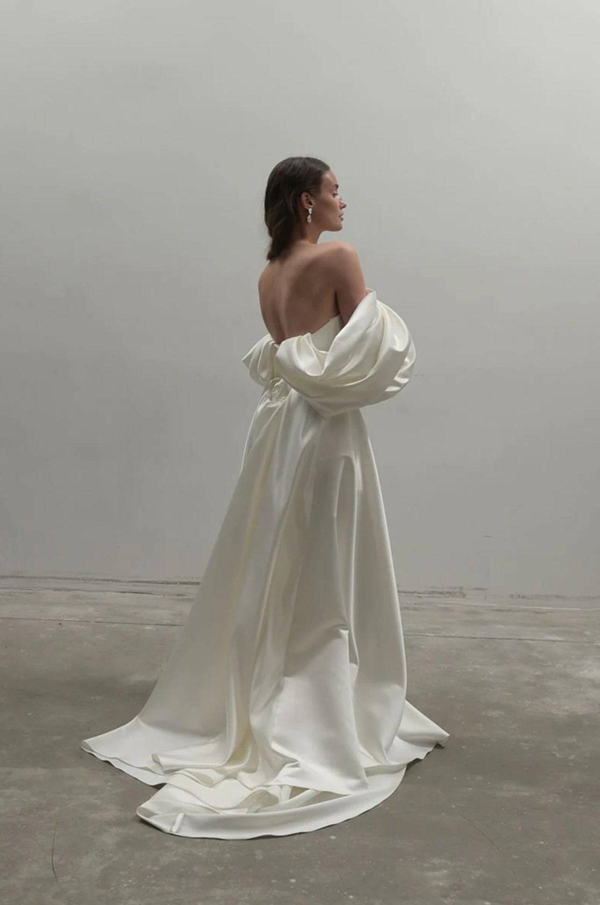 Off-shoulder white satin A-line gown with voluminous sleeves and a flowing train against a neutral background.
