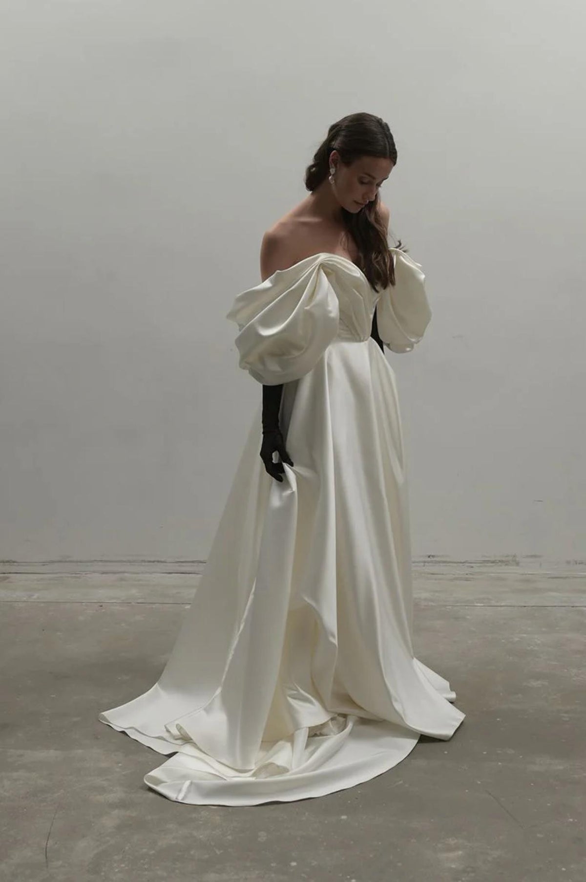 White off-shoulder satin A-line gown with voluminous sleeves, a pleated bodice, and a flowing floor-length skirt.