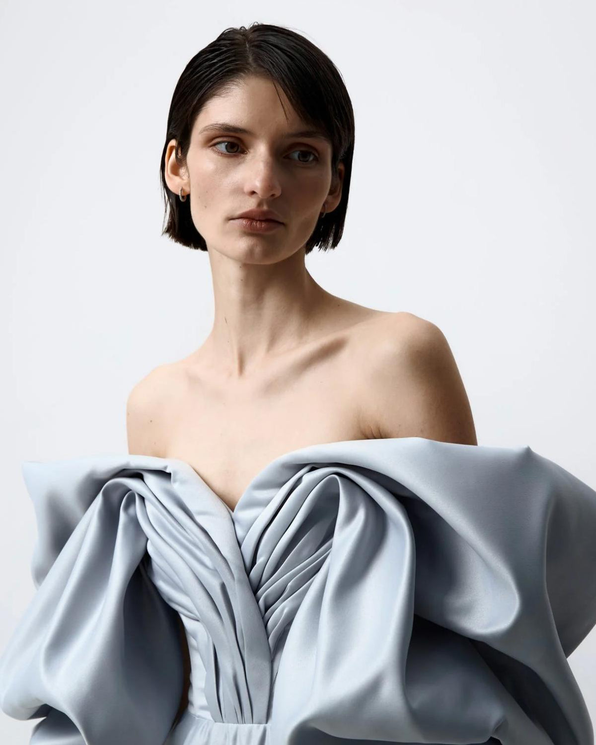 Model wearing a light blue, off-shoulder satin F1 gown with elegant, voluminous pleats and a structured bodice.