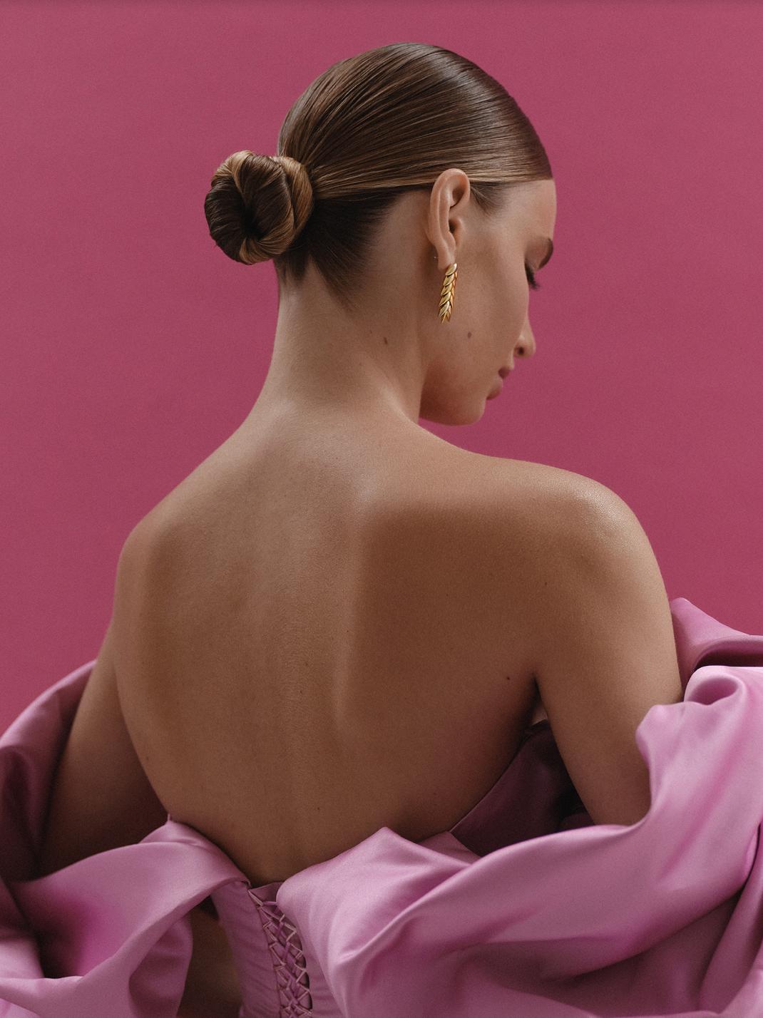 Woman wearing a pink off-shoulder satin F1 gown with a corset back, styled with a sleek bun and gold leaf earrings.
