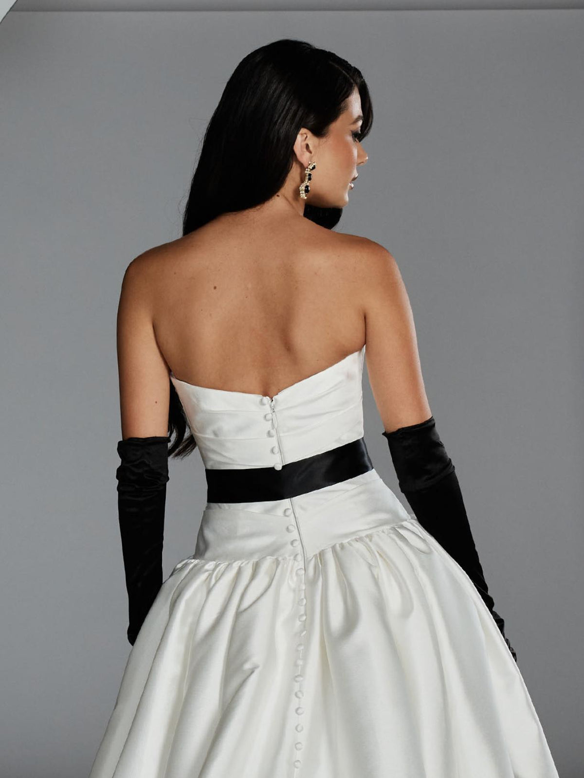 Back view of a strapless white Mikado ball gown with a black waist sash, button-down back, and long black opera gloves.