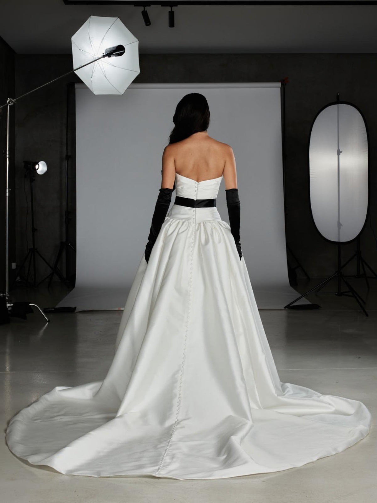 Back view of a white Mikado ball gown with a strapless bodice, black waist sash, long black gloves, and a button-up train.