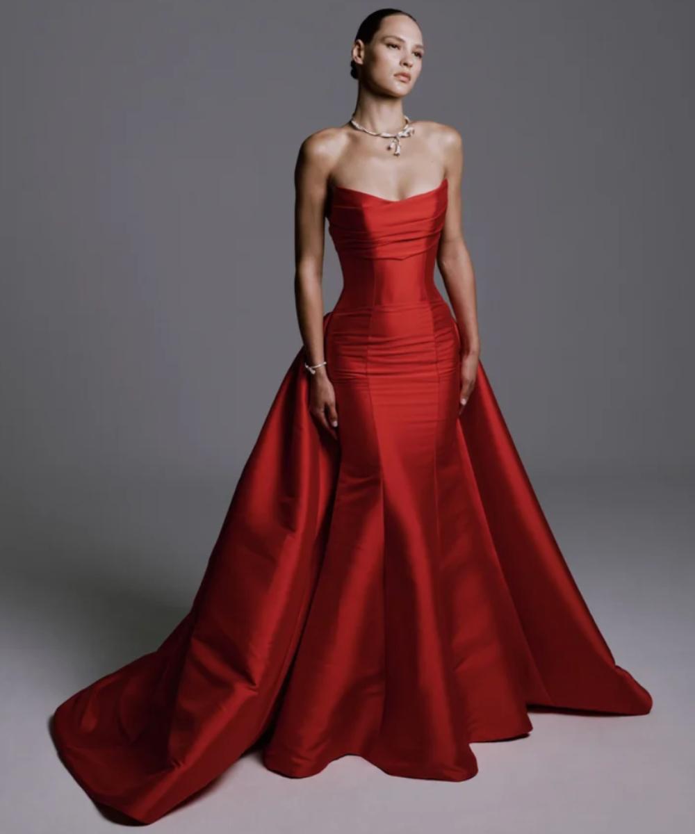 Model wearing a red strapless mikado gown with a structured bodice, sculptural overskirt, and dramatic train.