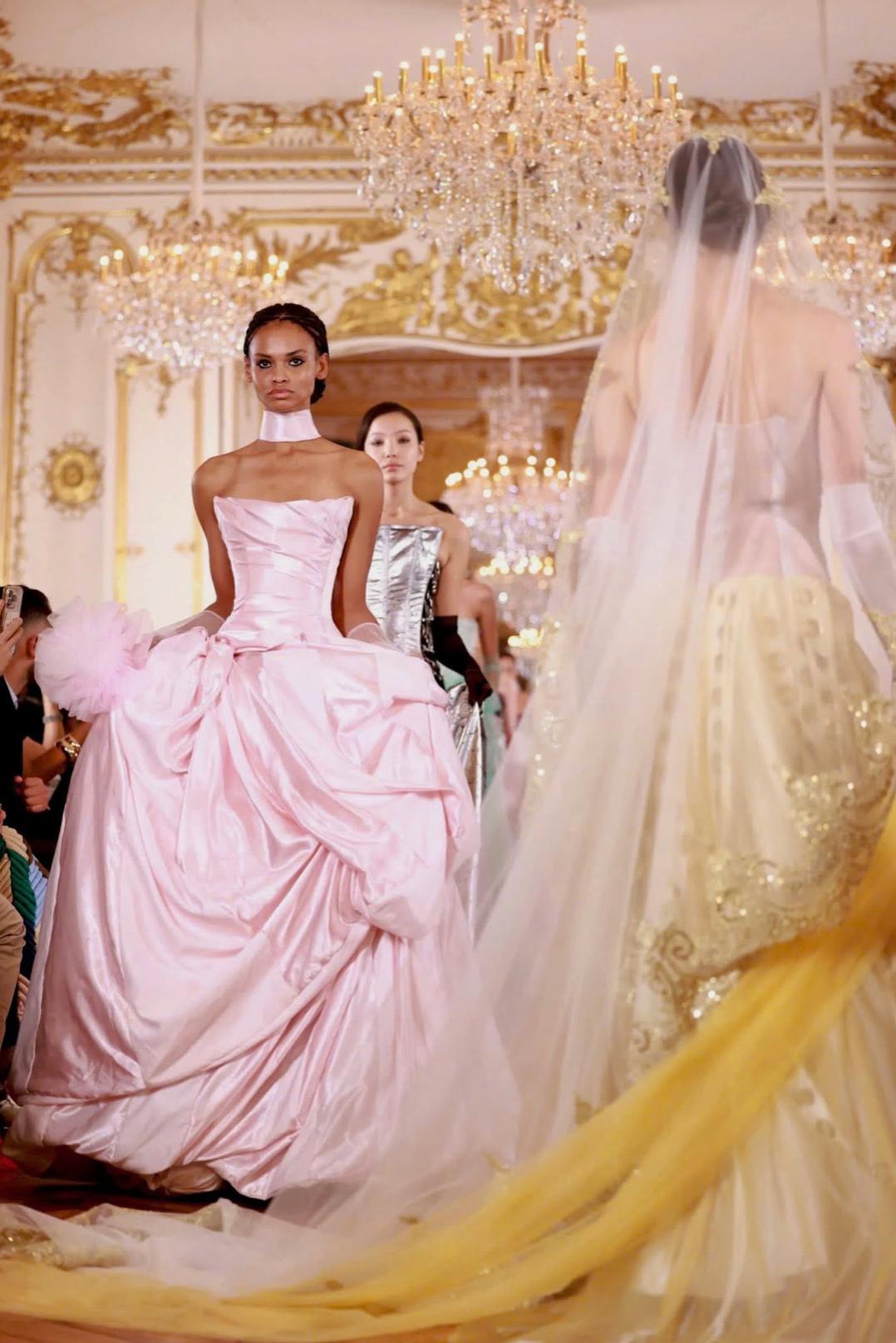Model wearing a pale pink off-shoulder satin A-line gown with a pleated bodice and voluminous skirt on a runway.