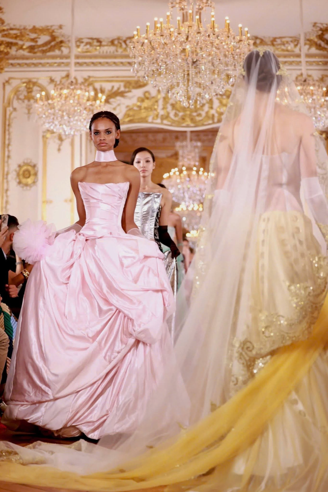 Model wearing a pale pink off-shoulder satin A-line gown with a pleated bodice and voluminous skirt on a runway.