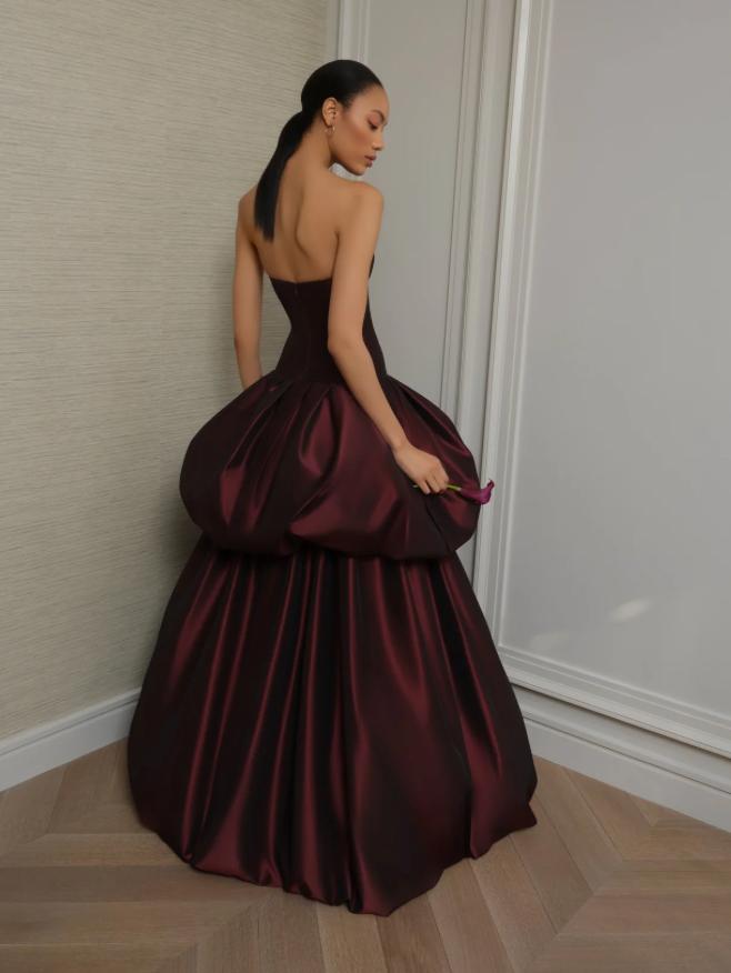 Strapless burgundy silk taffeta ball gown with a structured corseted bodice and a dramatic, voluminous tiered skirt.