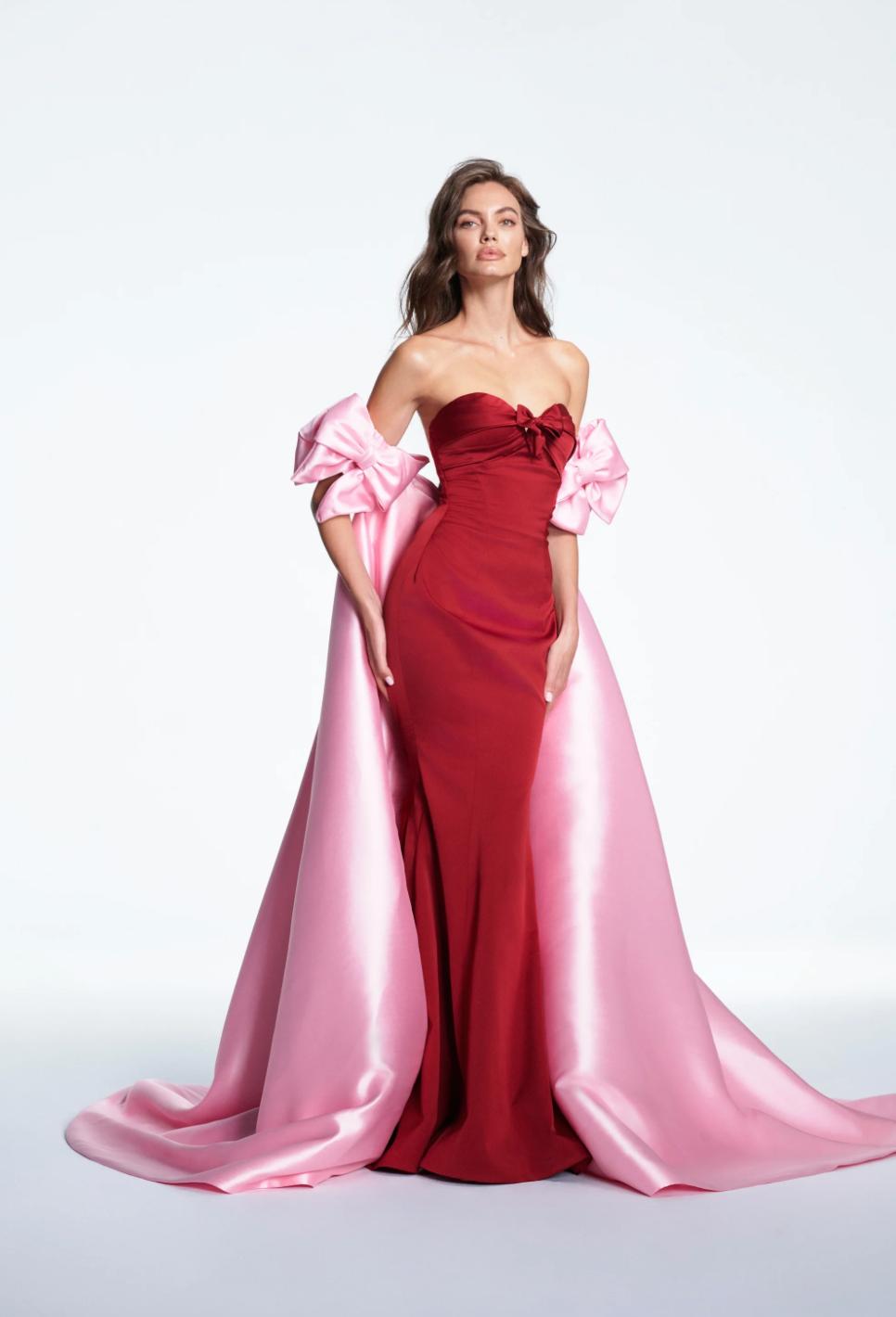 Red satin mermaid gown with a sweetheart neckline, featuring a dramatic pink cape and oversized bow details.