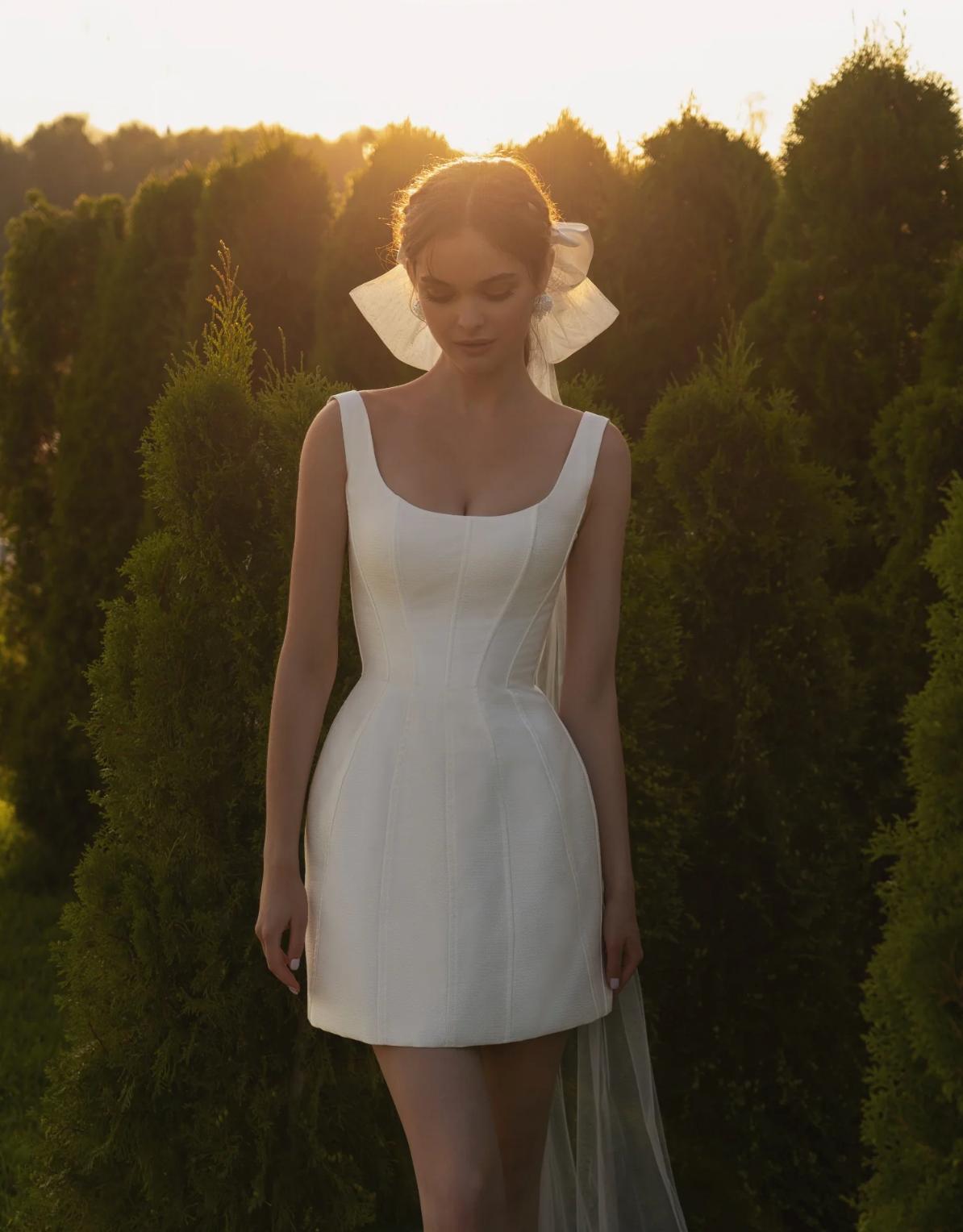 White structured bridal mini dress featuring a square neckline, corset bodice, and thin straps in a luminous fabric.