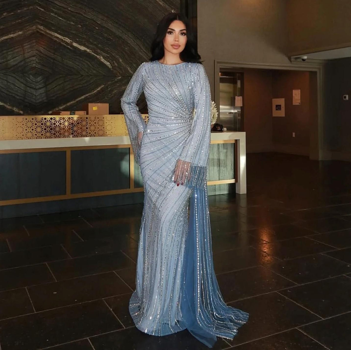 Floor-length light blue evening gown with sheer beaded sleeves, a fitted silhouette, and a dramatic side-skirt train.
