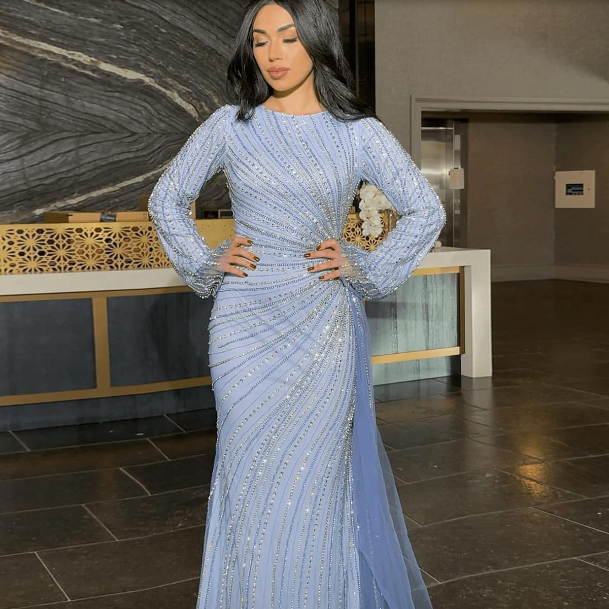 Light blue floor-length evening gown with long sheer sleeves, intricate silver beading, and a dramatic side-skirt detail.