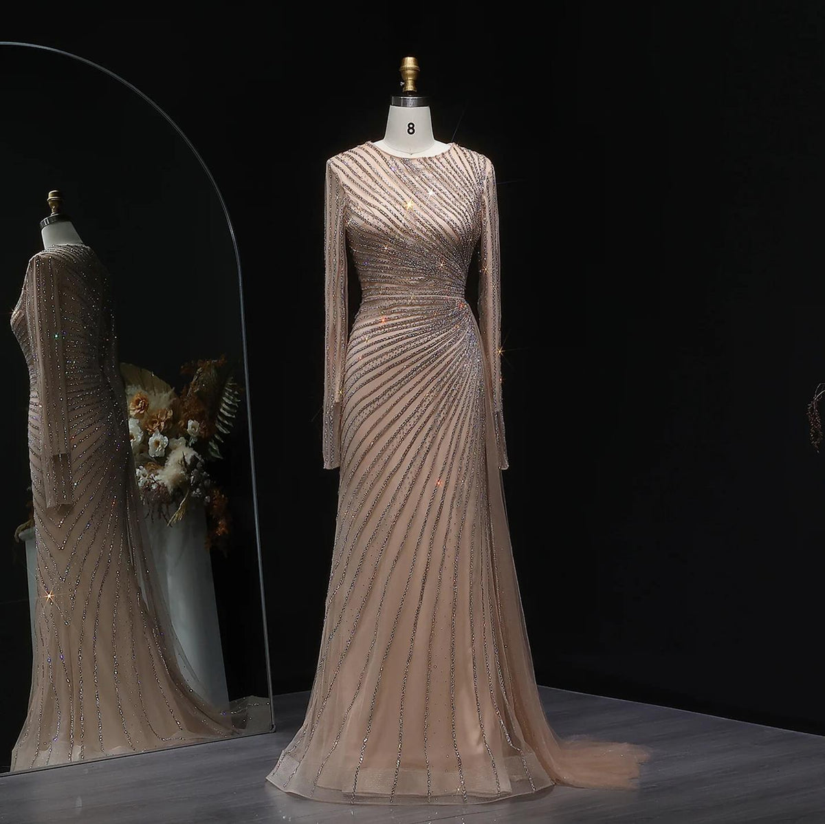 Beige floor-length evening gown with long sheer sleeves, intricate diagonal beading, and a sweeping train on a mannequin.